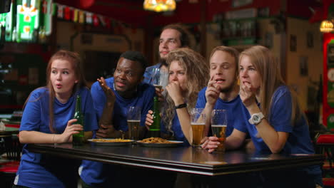 Un-Grupo-Multiétnico-De-Amigos-De-Aficionados-Con-Camisetas-Azules-Verán-Un-Partido-Por-Televisión-En-Un-Bar,-Verán-Un-Momento-Tenso-Y-Emocionante-Y-Celebrarán-Un-Gol-Marcado.-Abrazándonos-Divirtiéndonos-Y-Tomando-Un-Vaso-De-Cerveza.